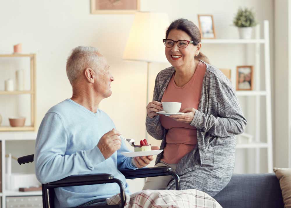 Mann im Rollstuhl isst mit seiner freundlichen Alltagsbegleitung Kuchen und lacht